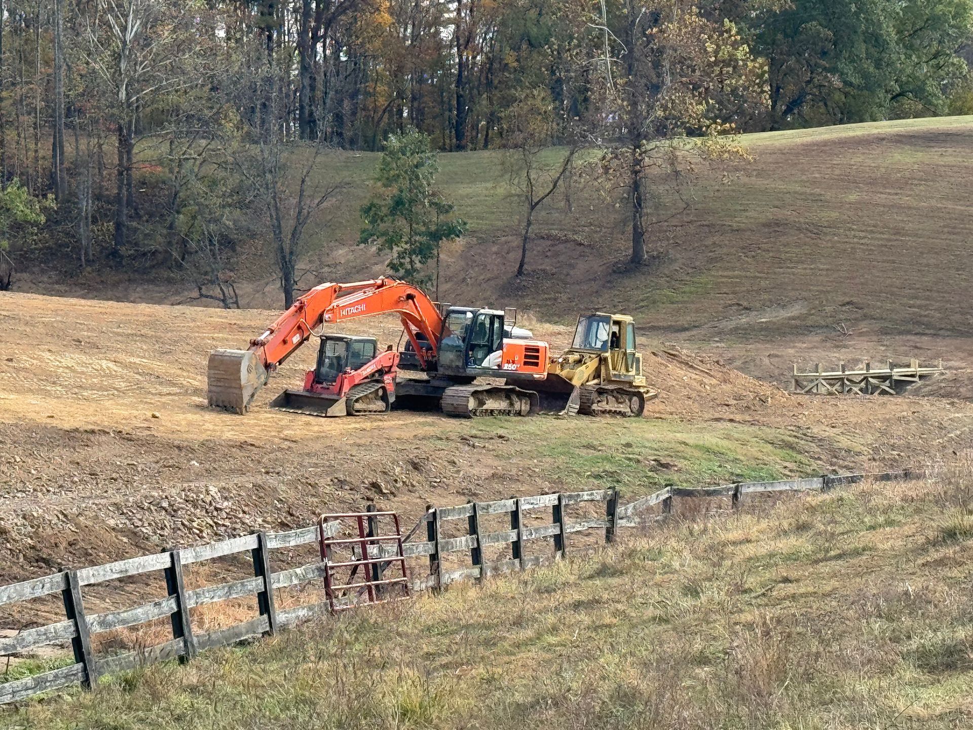 Construction equipment on a brown field near a wooden fence and trees; an orange excavator lifts a hay bale.