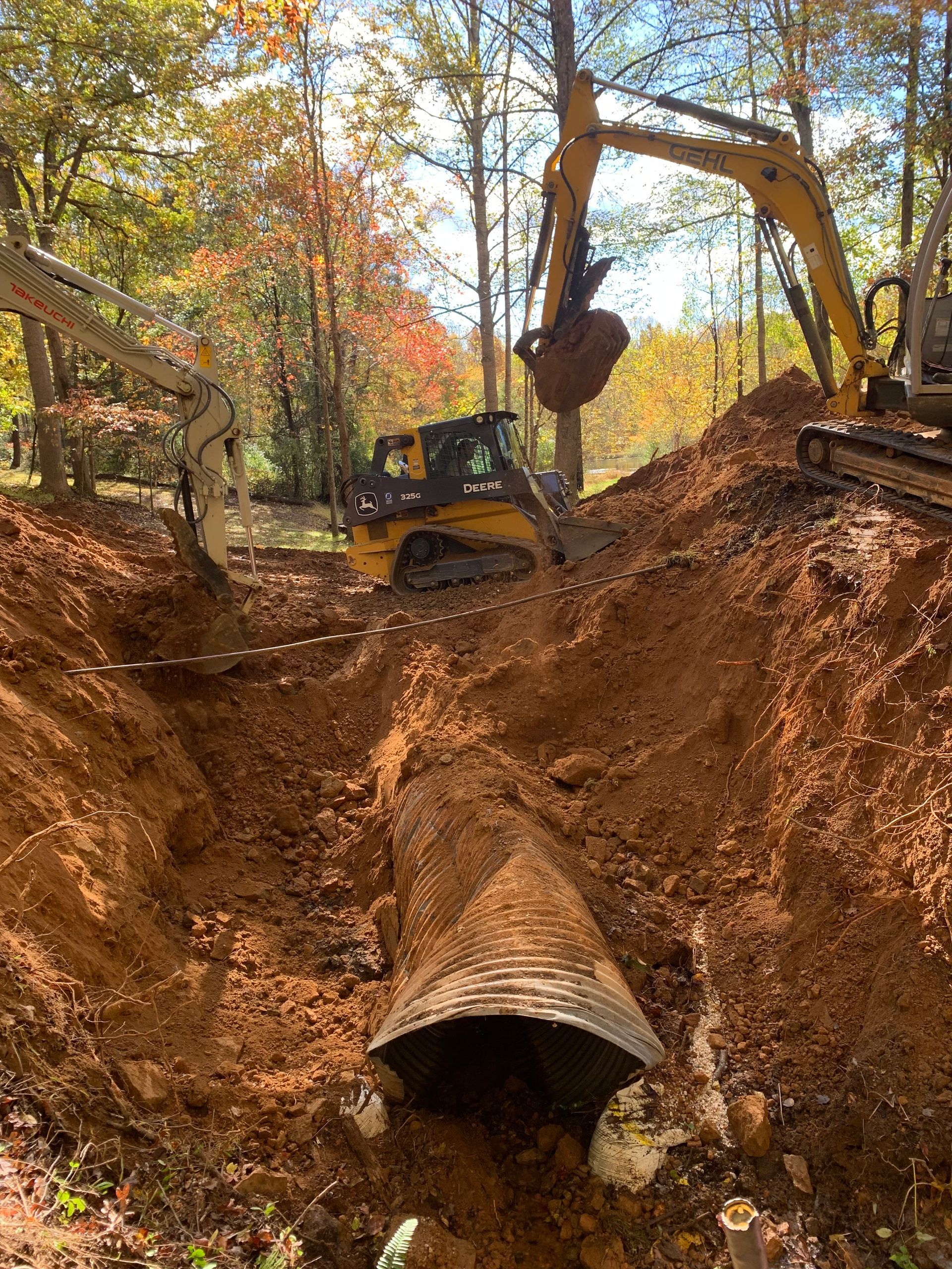 Construction site: excavators digging around a corrugated metal culvert in a forest.