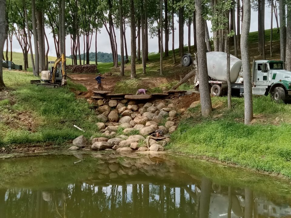 Construction site near a pond. Rock wall, trees, excavator, and cement truck visible. Two workers are present.