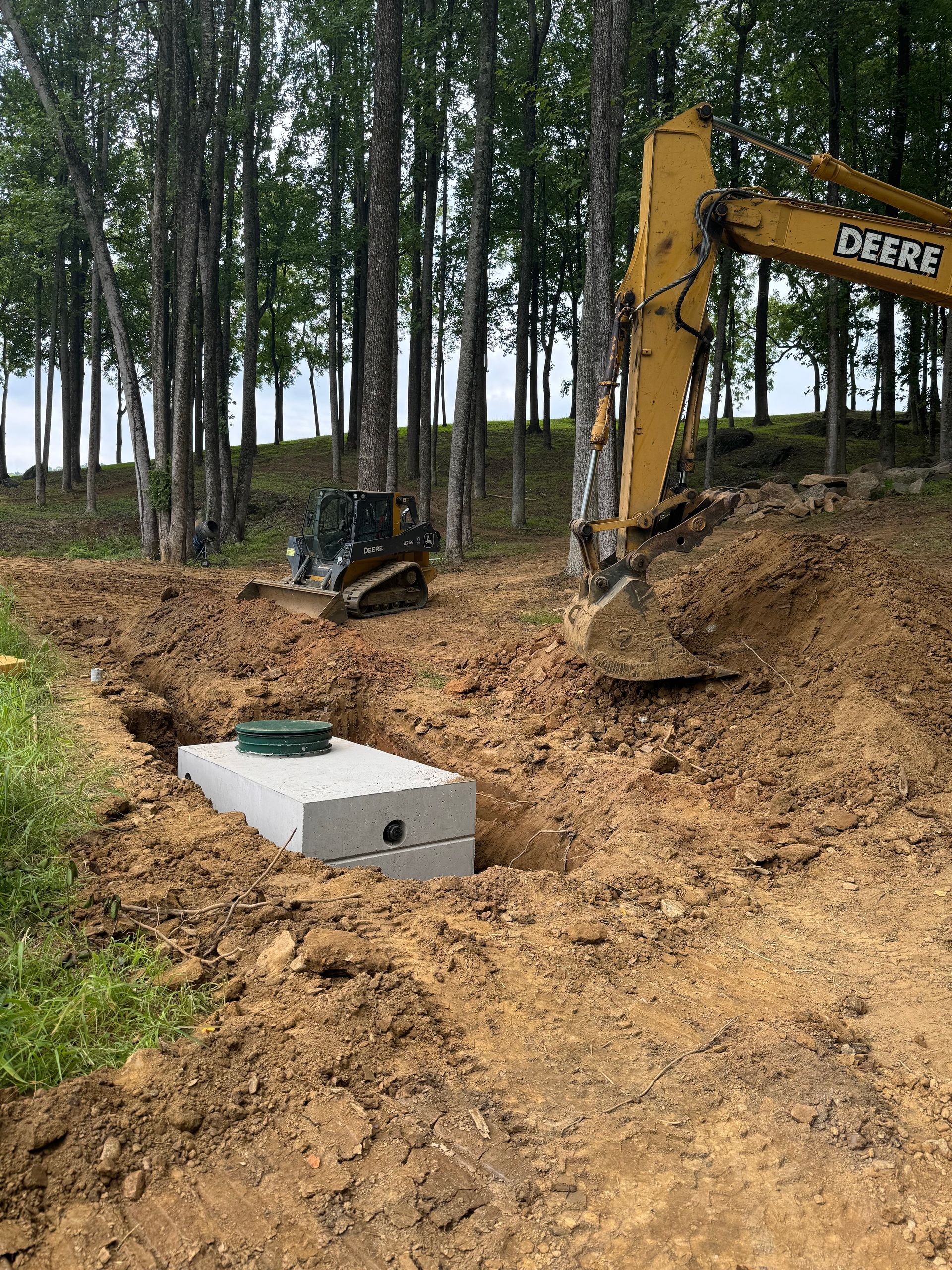 A gray concrete septic tank in an excavation pit with an excavator and a track loader in the background.