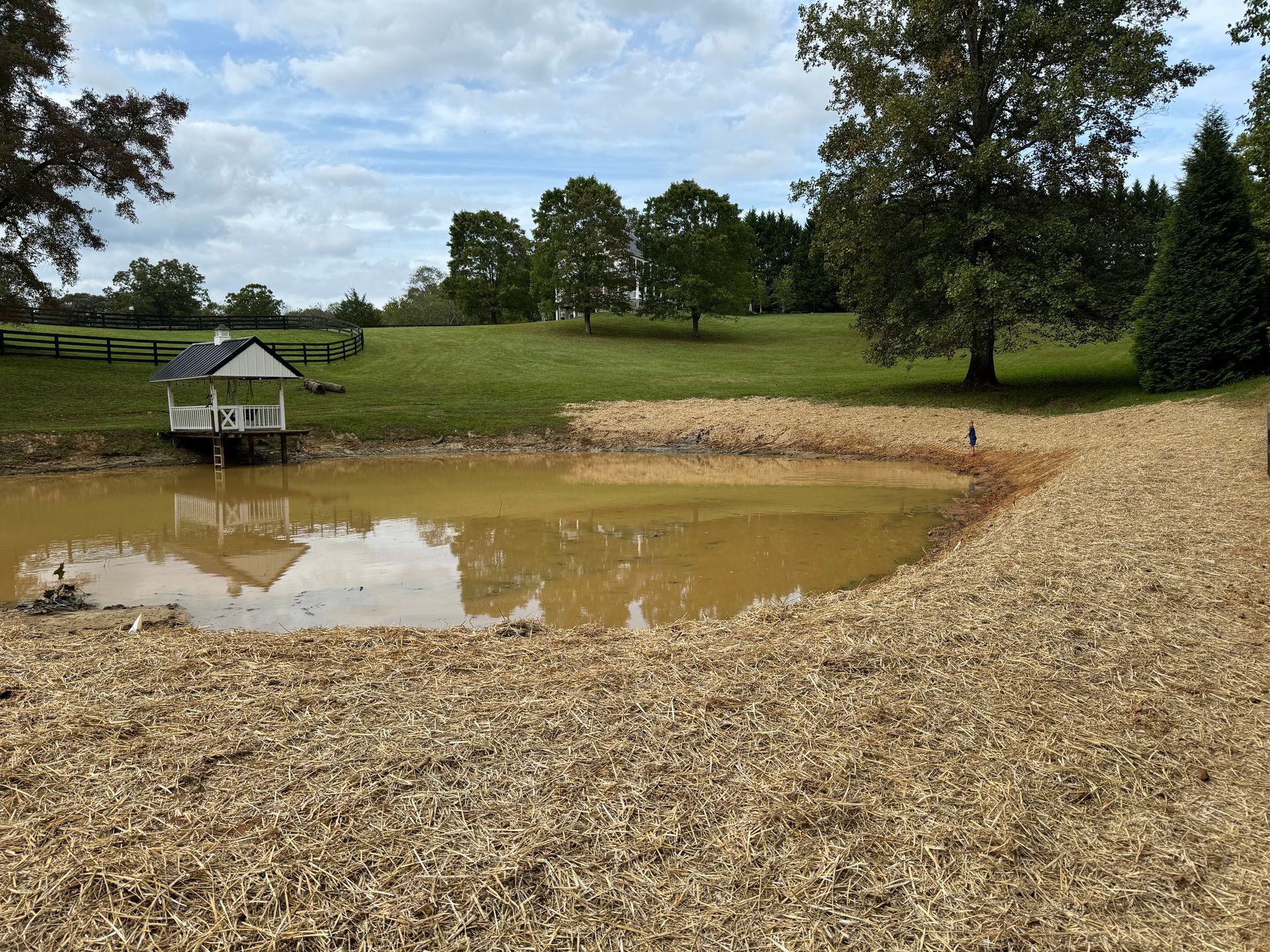 Muddy pond with wooden chip shoreline; a gazebo on a dock on the left, grassy field, and trees in the background.