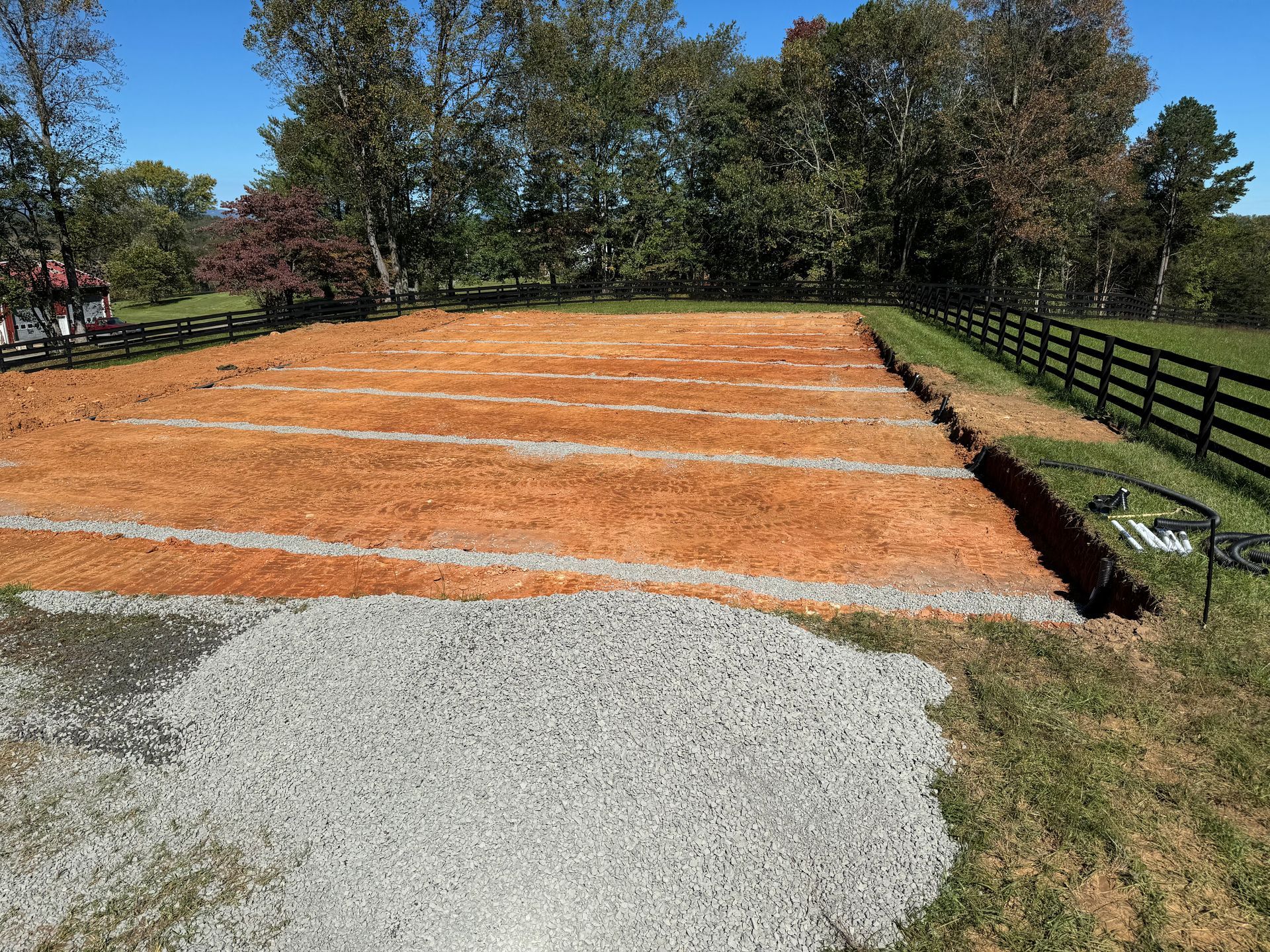 Construction site: red dirt foundation with gravel pile, white lines, and trench beside a black fence, blue sky.