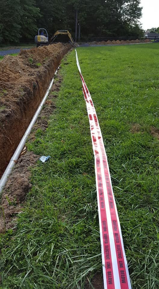 Trench dug in grass, with a white pipe and warning tape laid parallel. An excavator is in the background.