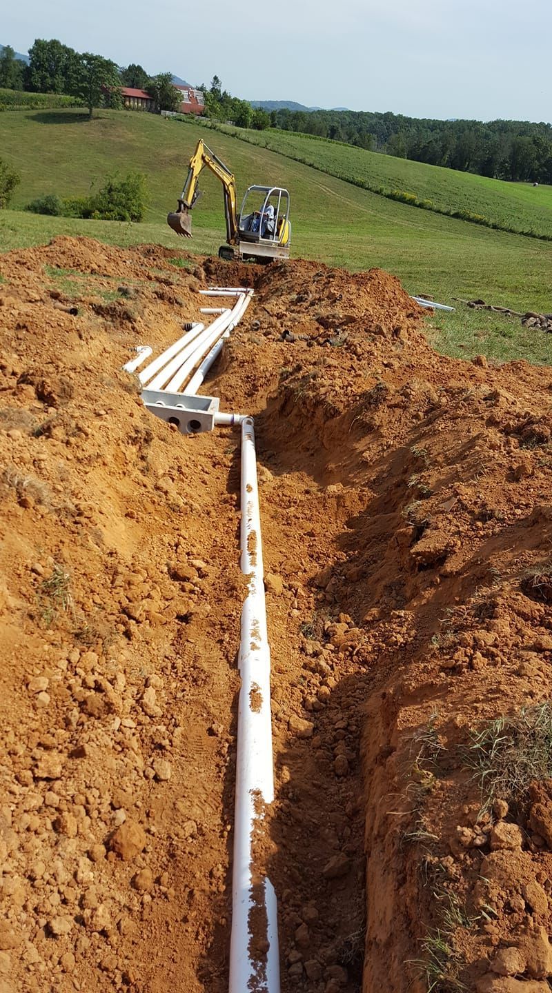 Trench with white pipes, backhoe in background, dirt and green hillside.