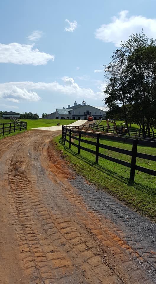Dirt driveway leading to a large white building with green fields and a wooden fence on a sunny day.