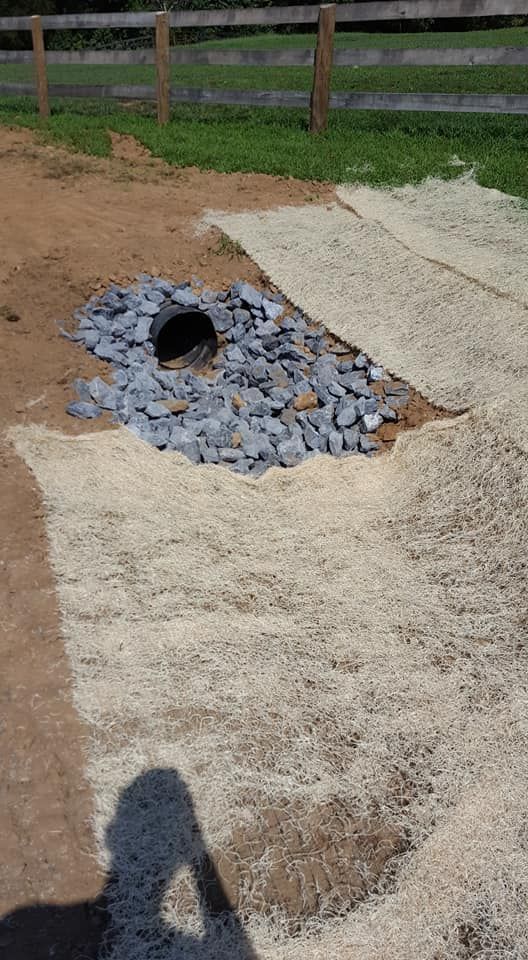 A drainage system with a black pipe surrounded by gravel, on a dirt and straw surface.