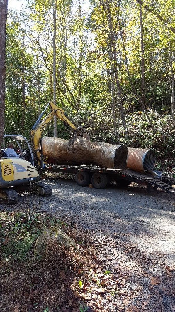 Excavator loading a large, cylindrical object onto a trailer on a gravel road in a wooded area.