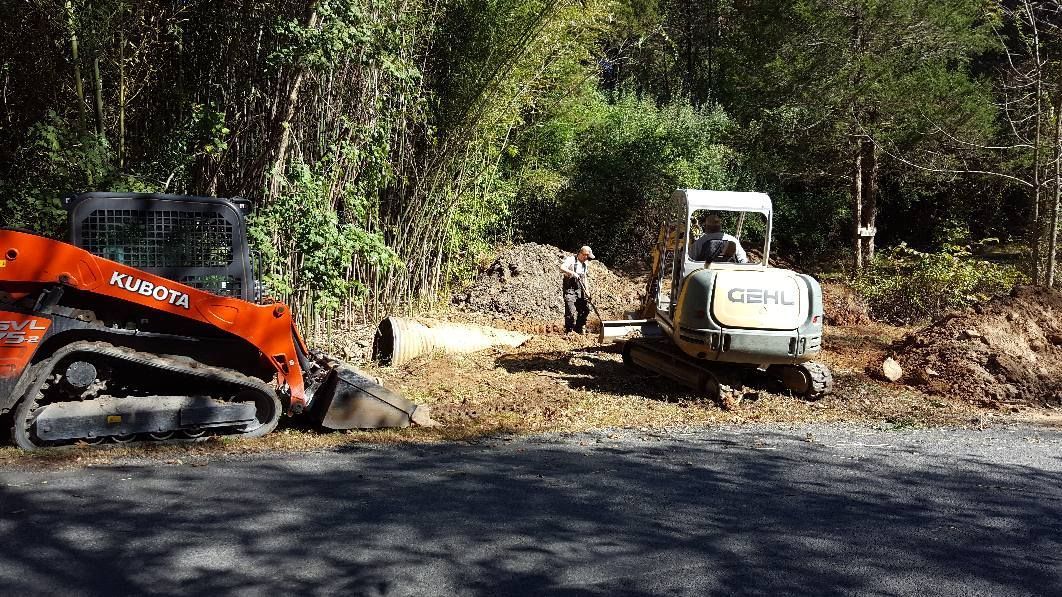 Orange skid steer and small excavator at a construction site with a person standing nearby; forest backdrop.