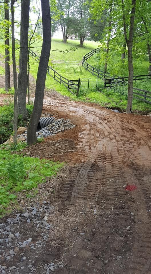 Dirt road crossing a culvert, leading uphill to a fence and green hillside with trees on either side.