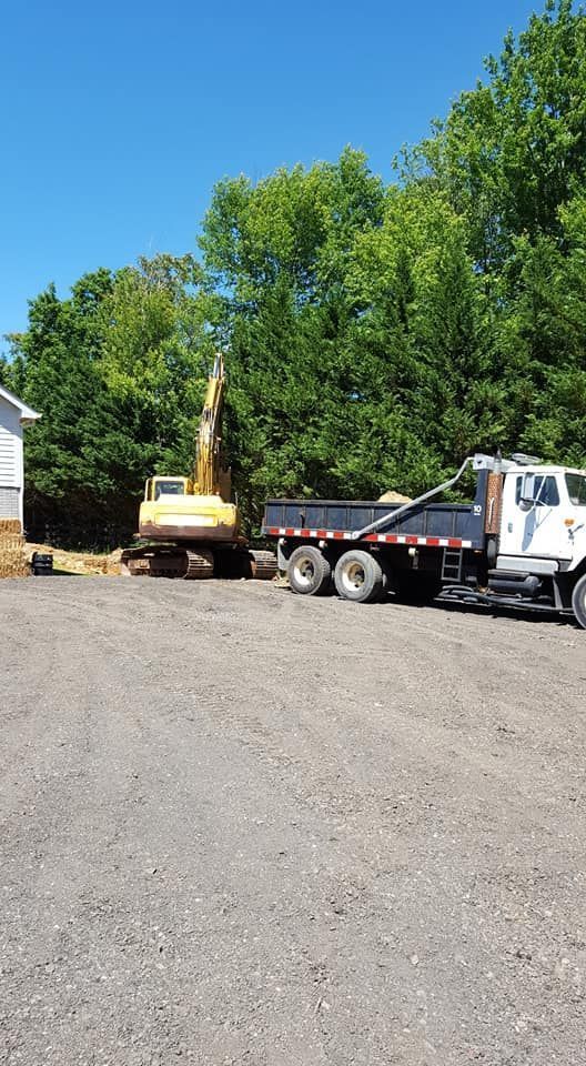 Yellow excavator loading a dump truck at a construction site, trees in the background, blue sky.