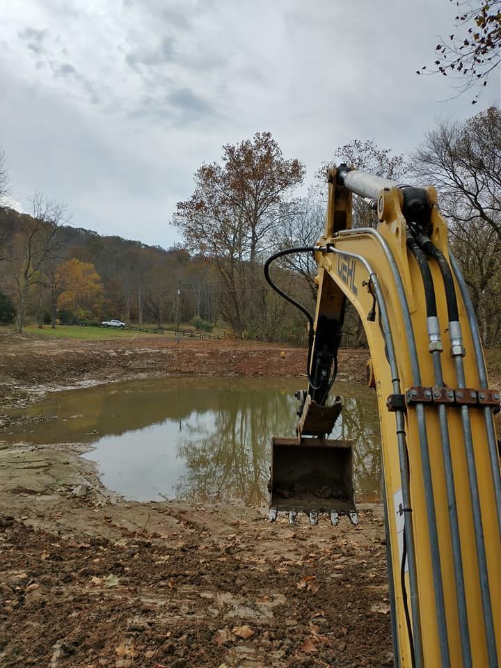 An excavator bucket next to a pond in a wooded area under an overcast sky.