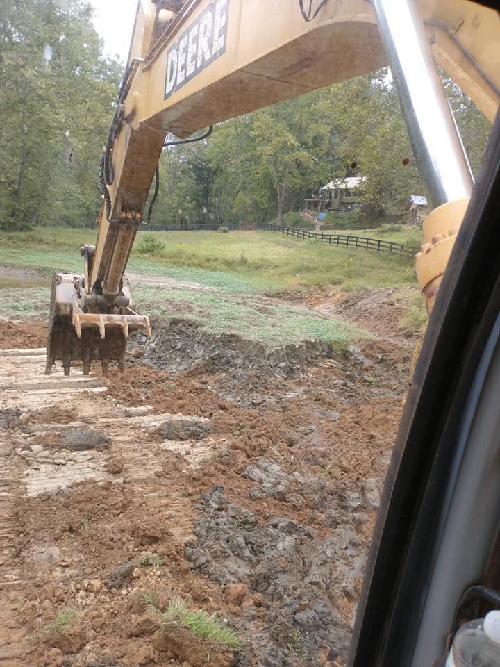 Yellow Deere excavator digging in brown dirt, with green trees and a building in the background.