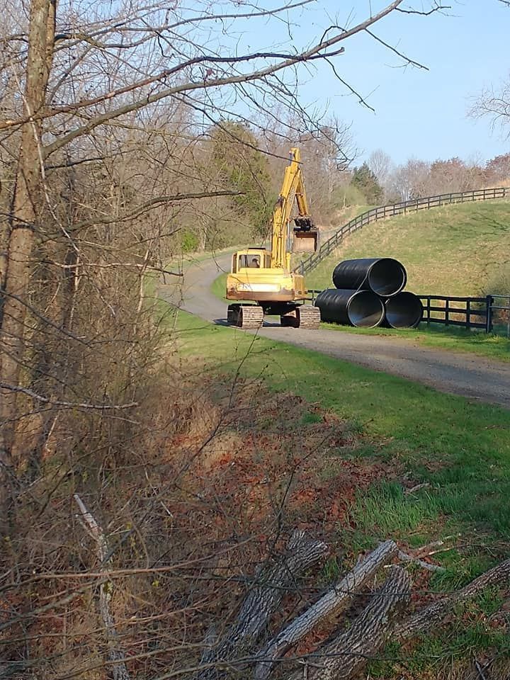 Yellow excavator on a dirt road next to large black pipes. Rolling green hills in the background.