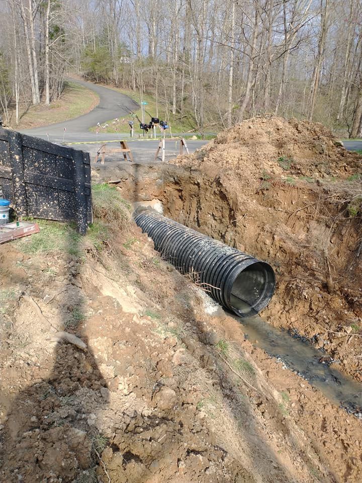 Black corrugated drainage pipe in a deep trench. Road and trees in the background, dirt pile on the right.