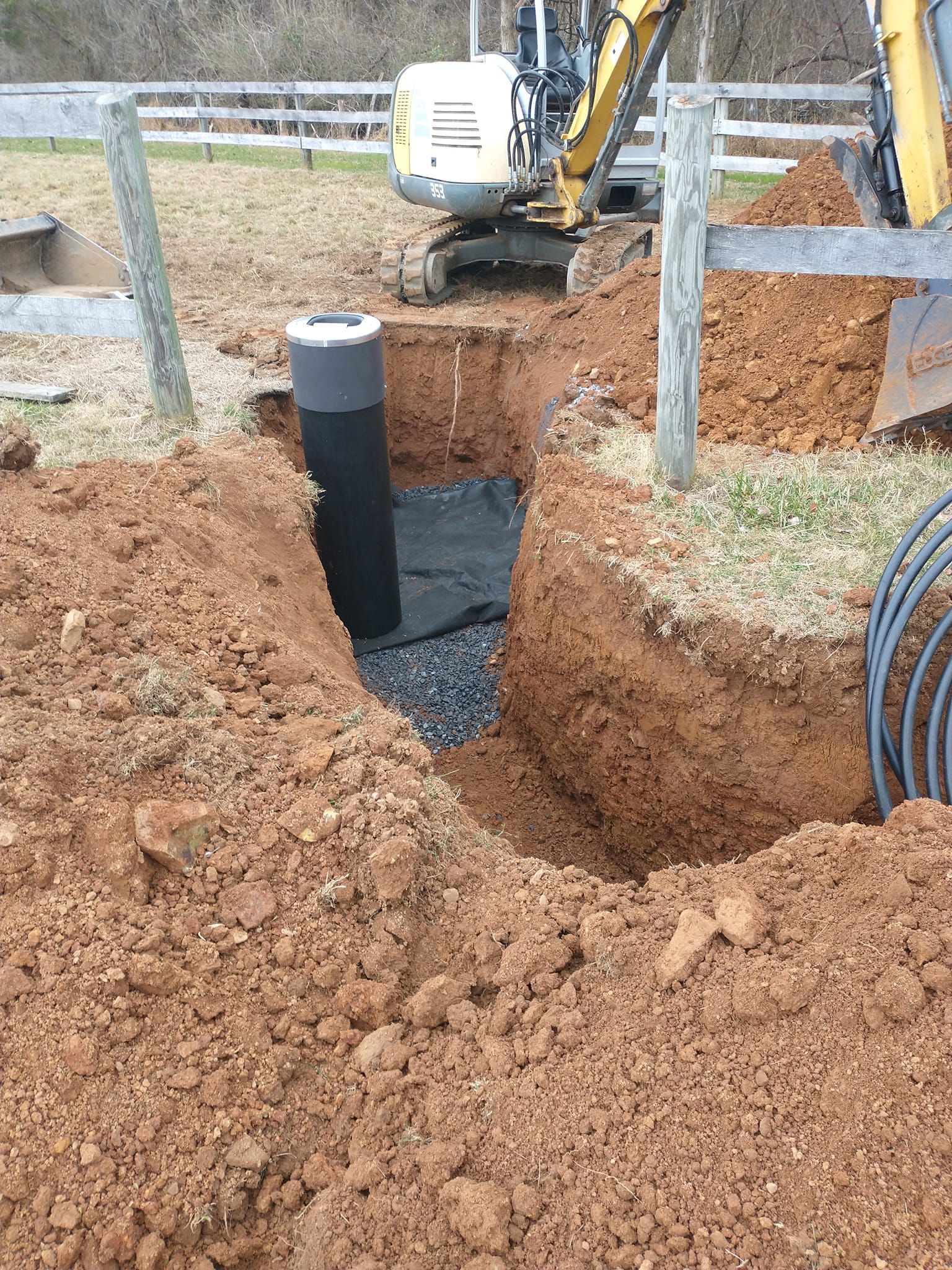 Excavation site with an excavator, black pipe, and gravel. Earth and fence posts are visible.