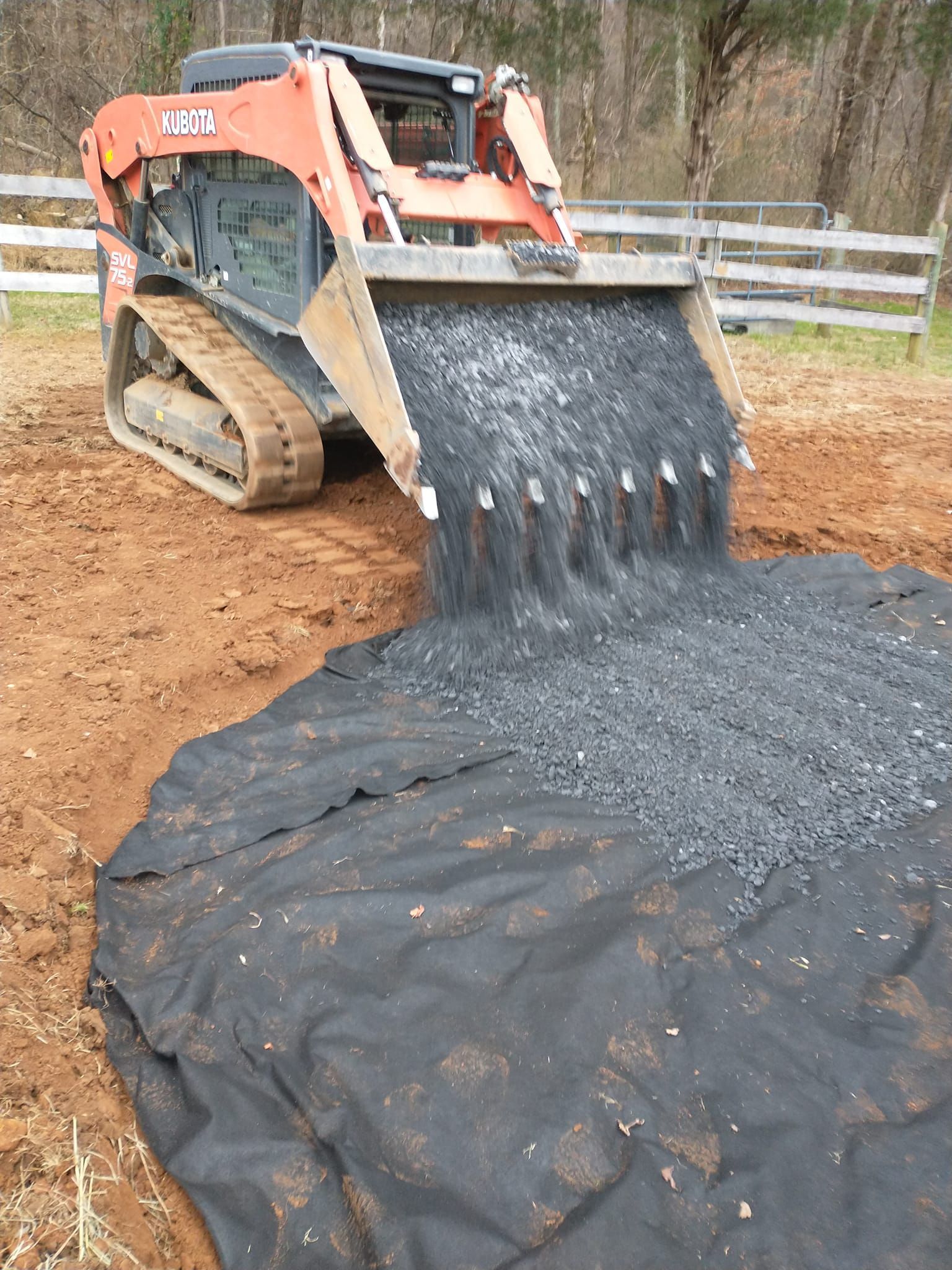 Skid steer dumping gravel onto a black fabric in a dirt trench, near a fence.