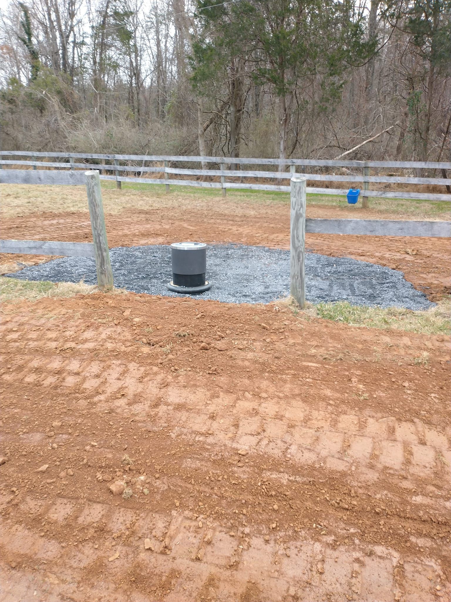 A cylindrical well cap on a gravel bed, within a wooden fence, surrounded by red dirt.