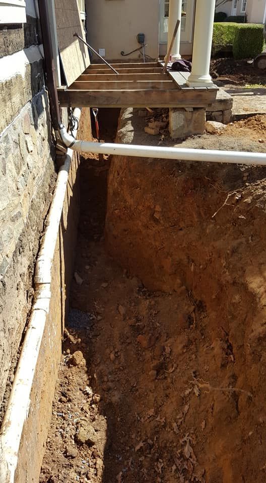 Trench dug along a building's foundation, with white pipes visible. Bare wood supports a porch.