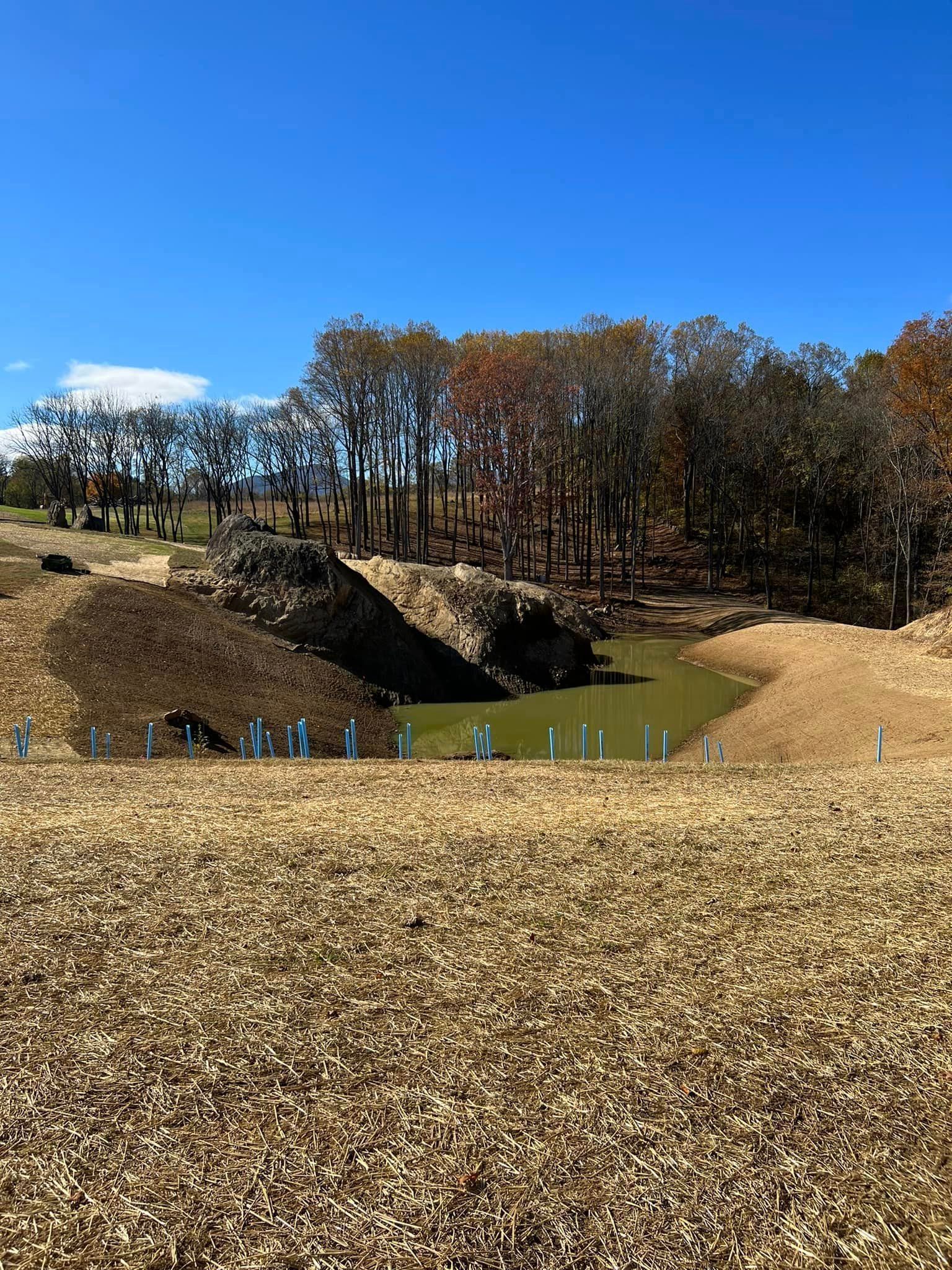 Construction site with water-filled pit, earth-colored landscape, and trees under a blue sky.