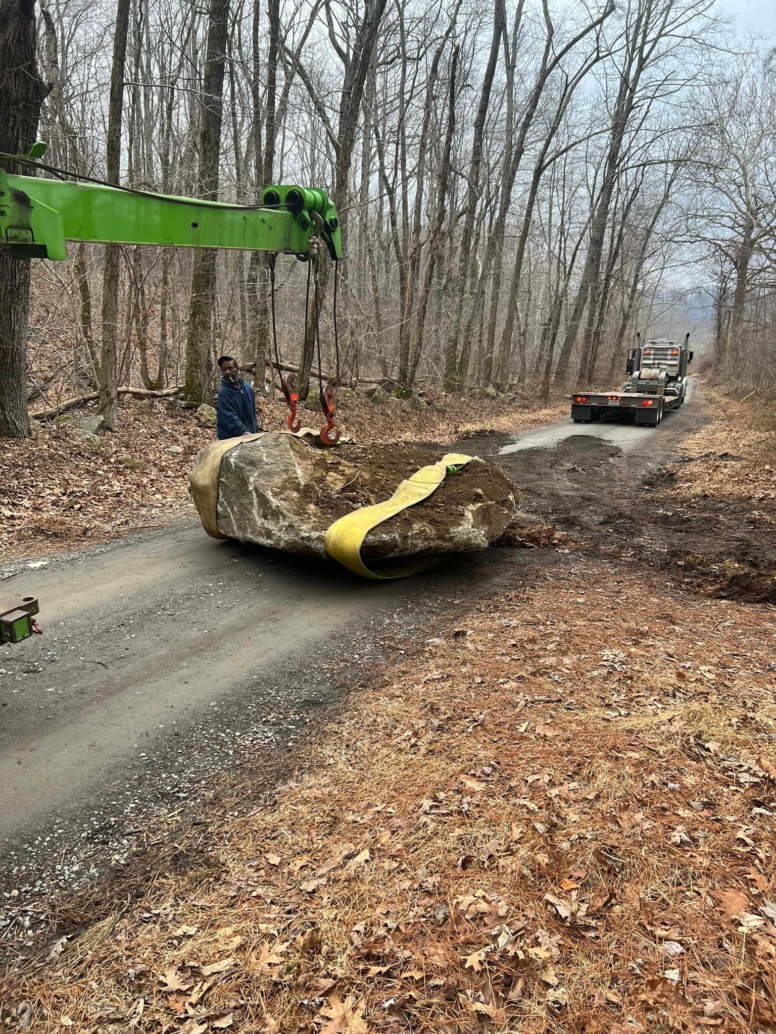 A large rock is being lifted off a dirt road by a green crane. A person stands nearby.