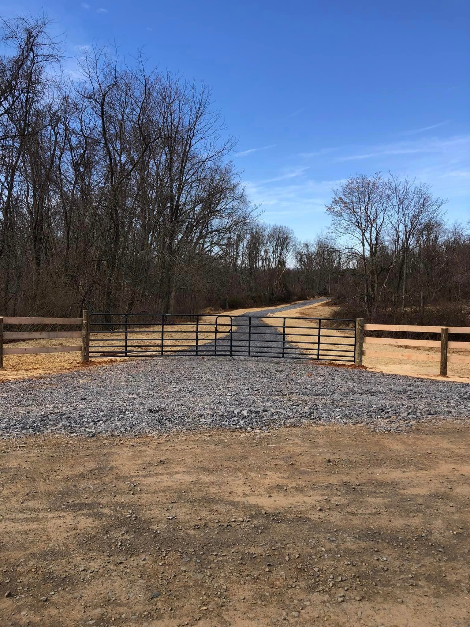Gravel path leads through a gate and fence into a wooded area on a sunny day.