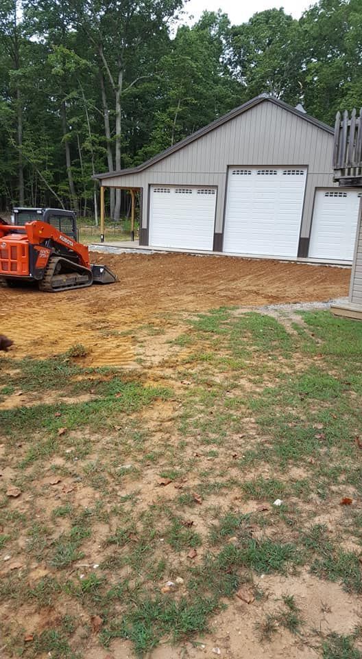 Orange skid steer excavating dirt in front of a gray garage with white doors, with green grass in the foreground.