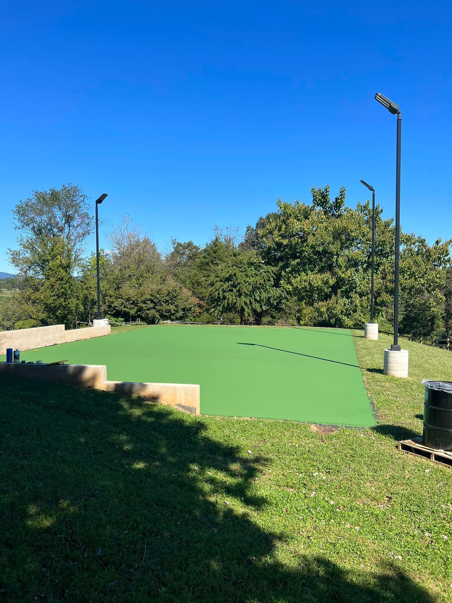 Green sport court with lighting and surrounding foliage under a clear, blue sky.