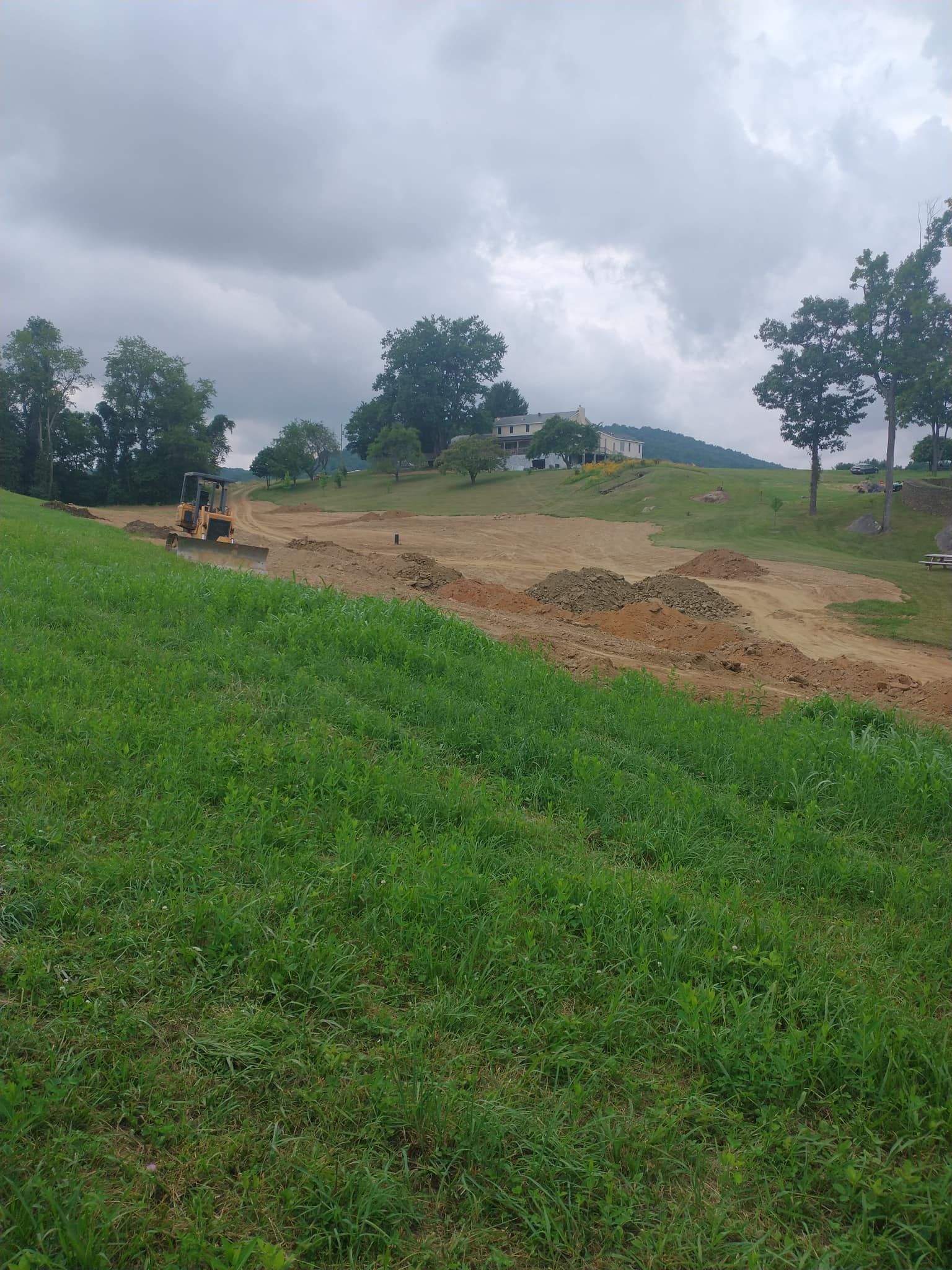 Green grassy hillside with a construction site, heavy equipment, and a distant house under a cloudy sky.
