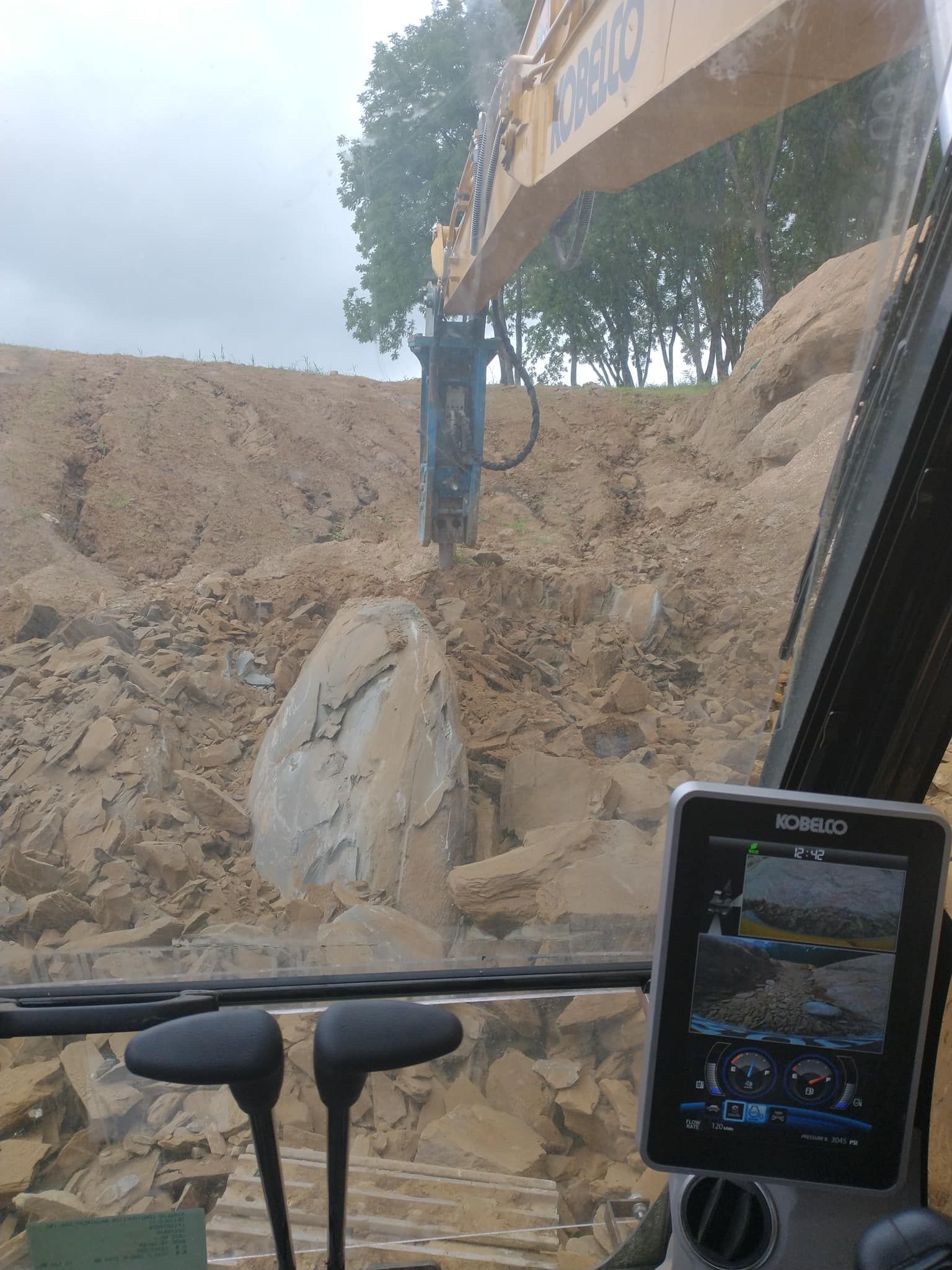 Excavator breaking a large rock, viewed from inside the cab. Brown earth surrounds the machine.