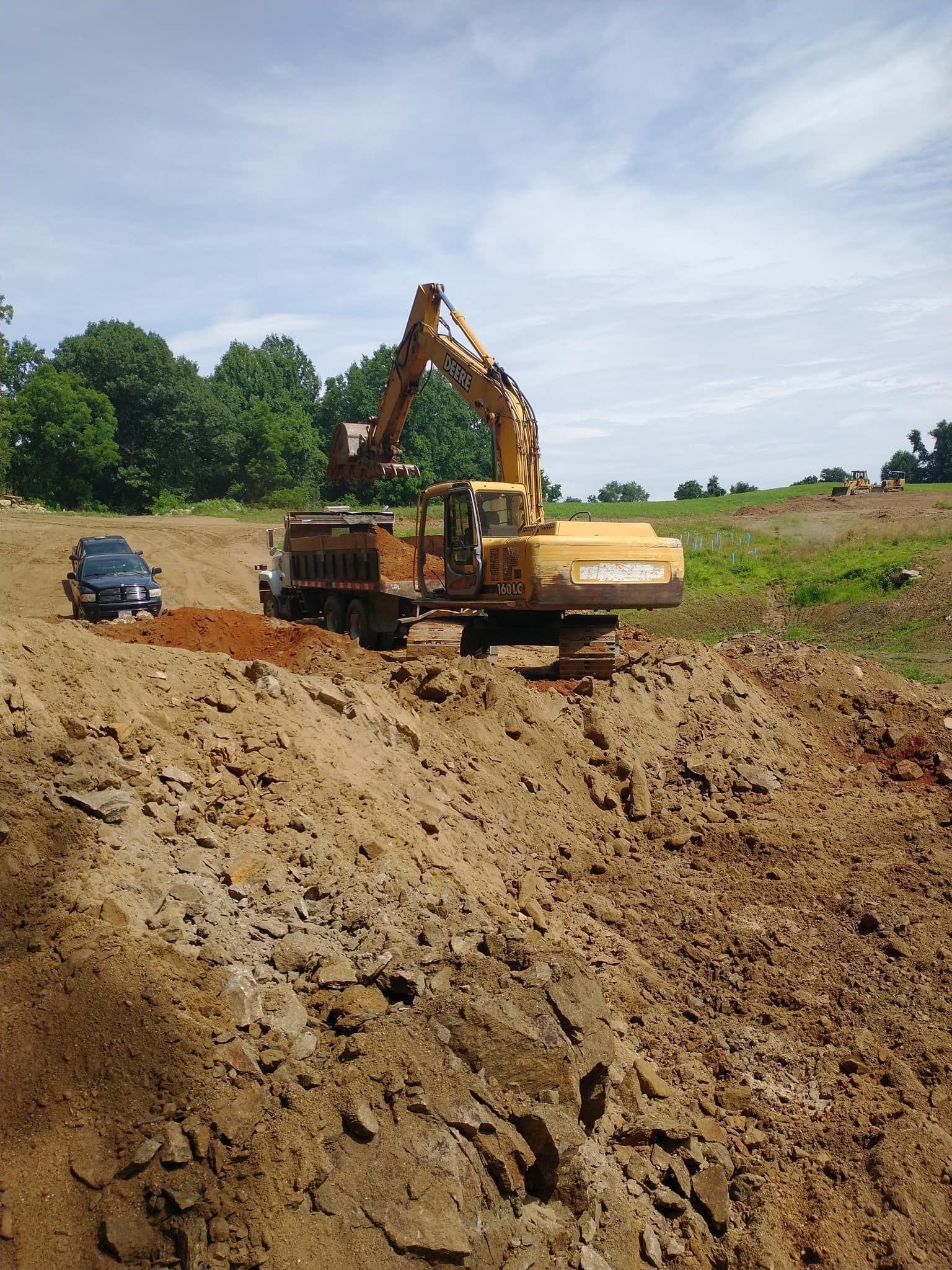 Excavator loading a dump truck with dirt on a construction site; overcast sky, trees in the background.