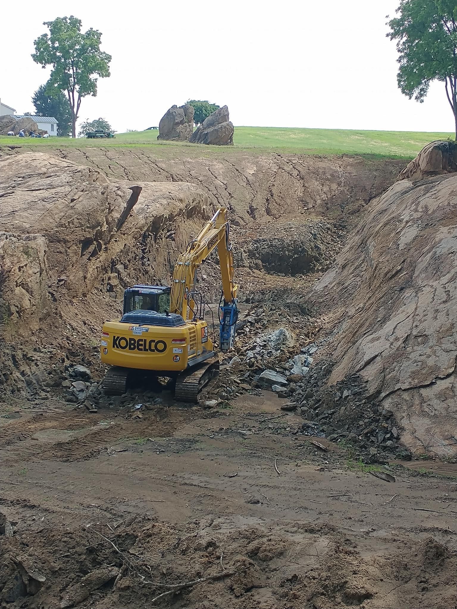 Yellow excavator in a deep trench, digging into the earth. Green grass and trees in the background.
