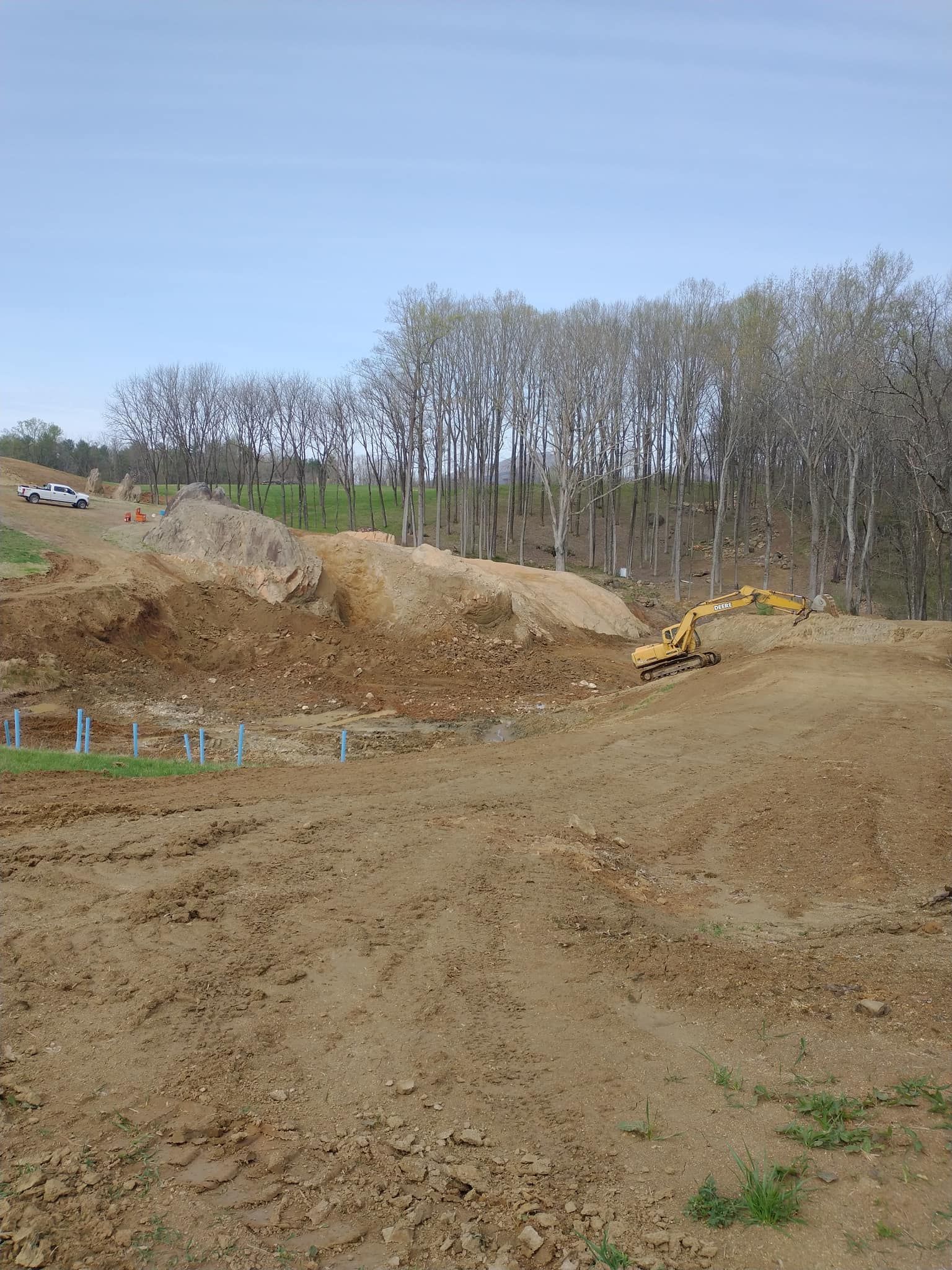 Construction site with excavator and dirt piles, trees in background, blue sky.