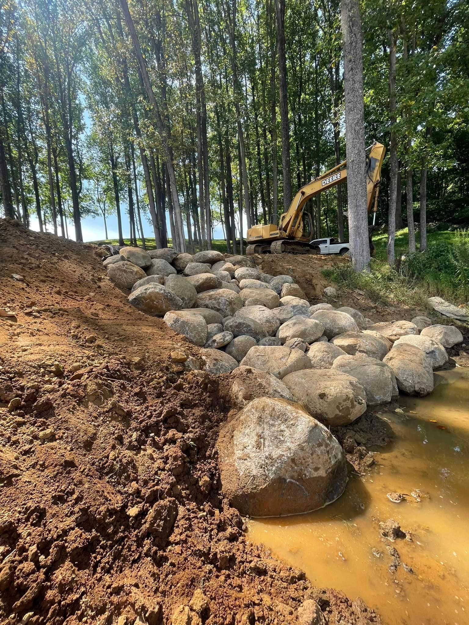 Construction site with large rocks and excavator near trees, muddy water, and exposed dirt.
