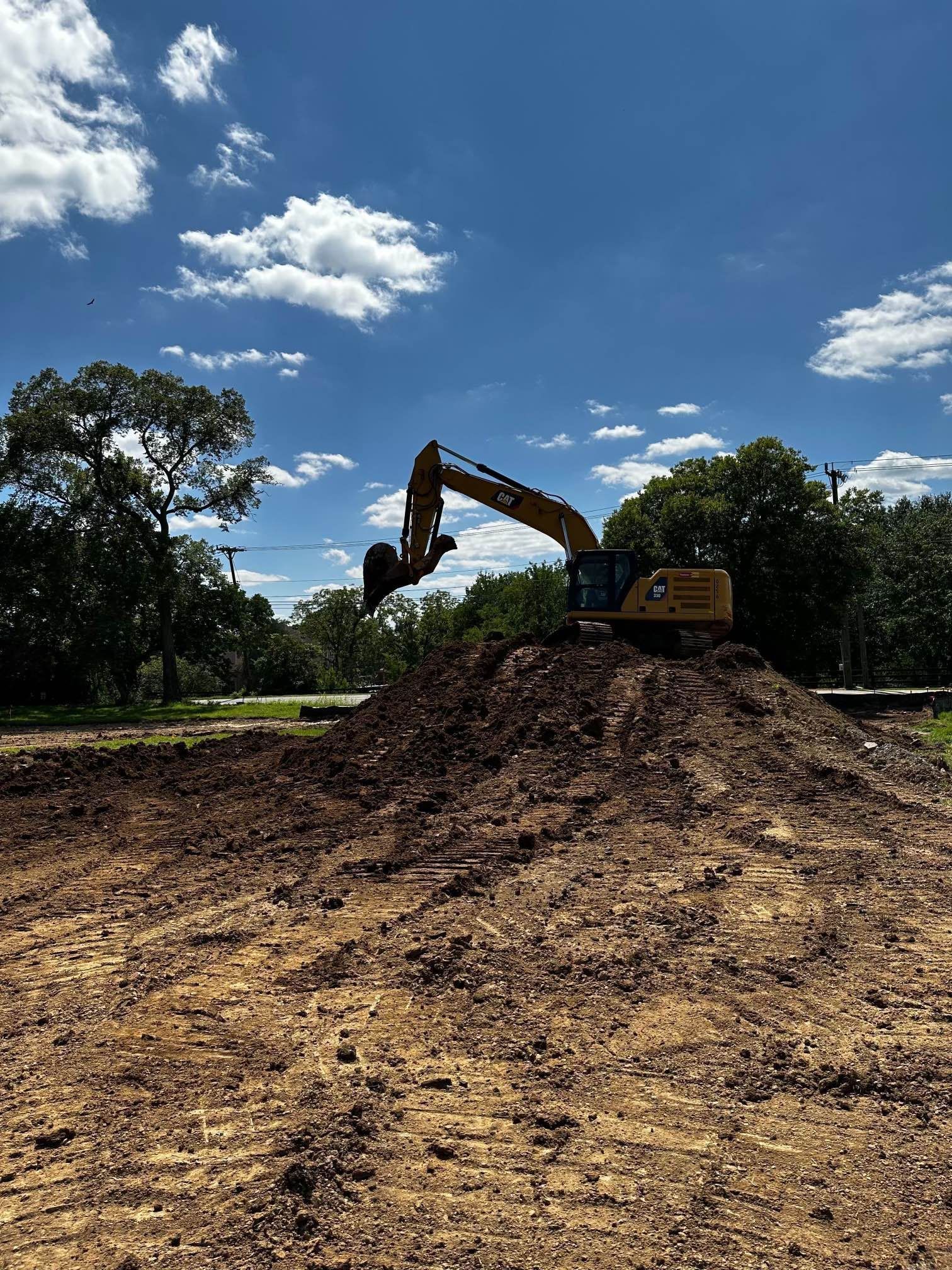 Excavator on a mound of dirt under a blue sky with clouds, trees in the background.