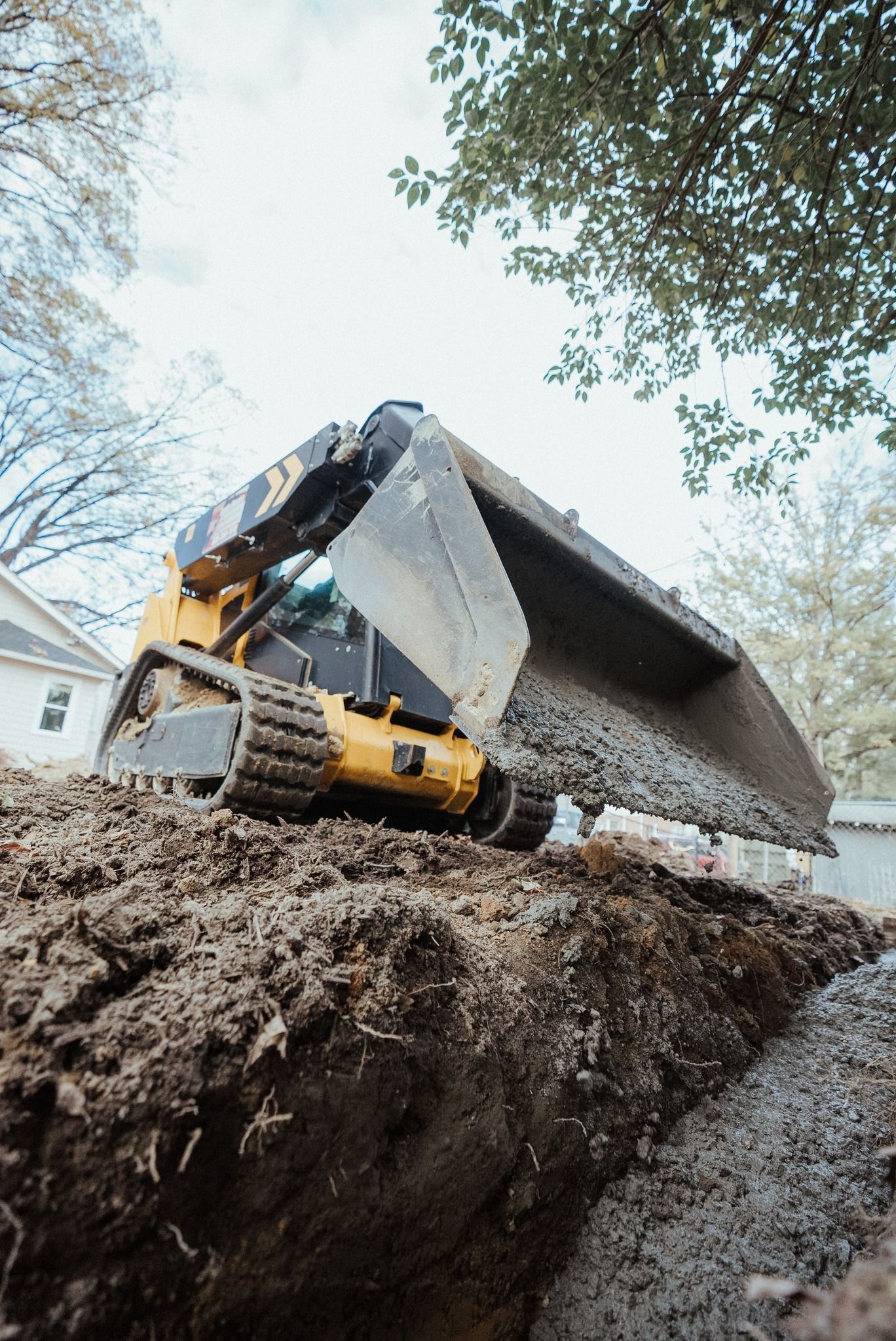 Yellow skid steer dumping gravel into a trench in a yard.