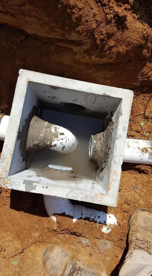 Concrete catch basin with white pipes, sitting in a trench in the soil.