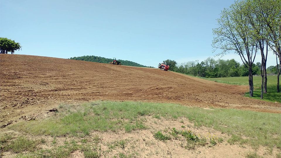 Dirt hill being leveled by tractors under a clear blue sky, with grass and trees in the foreground.