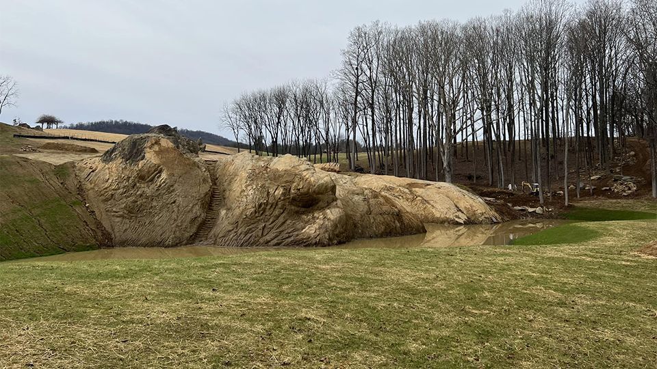 Large pile of straw on grassy field with bare trees in background. Cloudy sky.