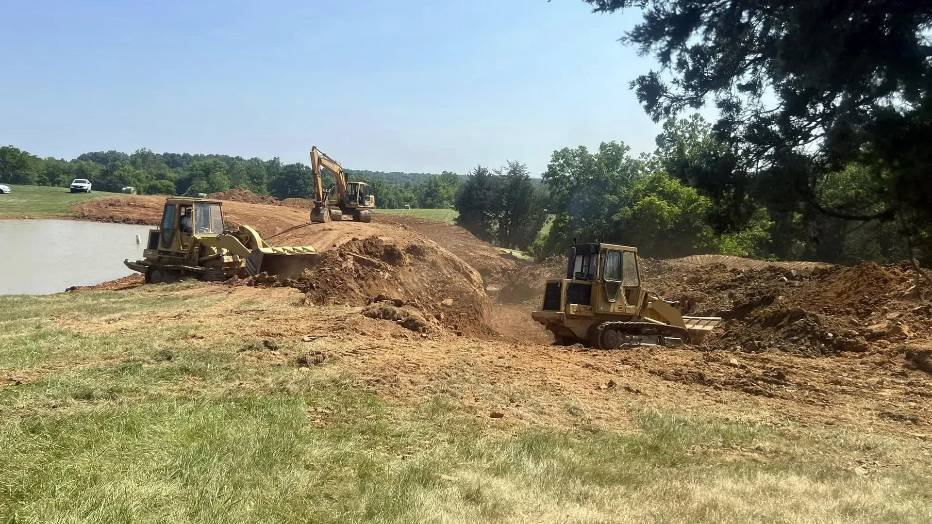 Construction site: Excavators moving earth near a body of water under a sunny sky.