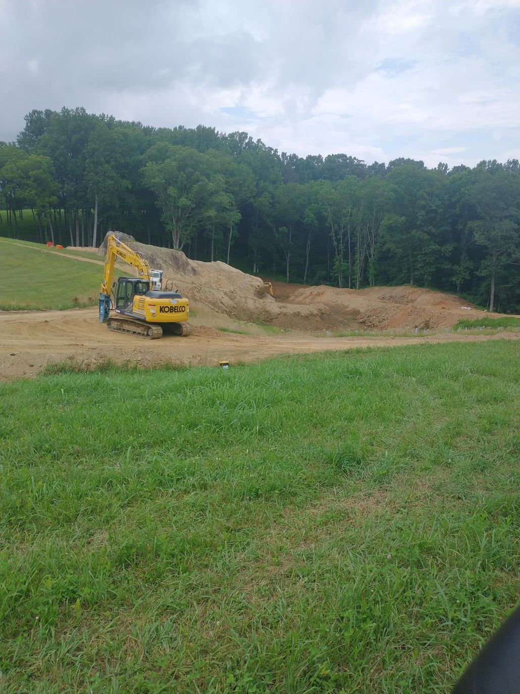 Yellow excavator on a dirt pile, working on a construction site next to a grassy field and a forest.
