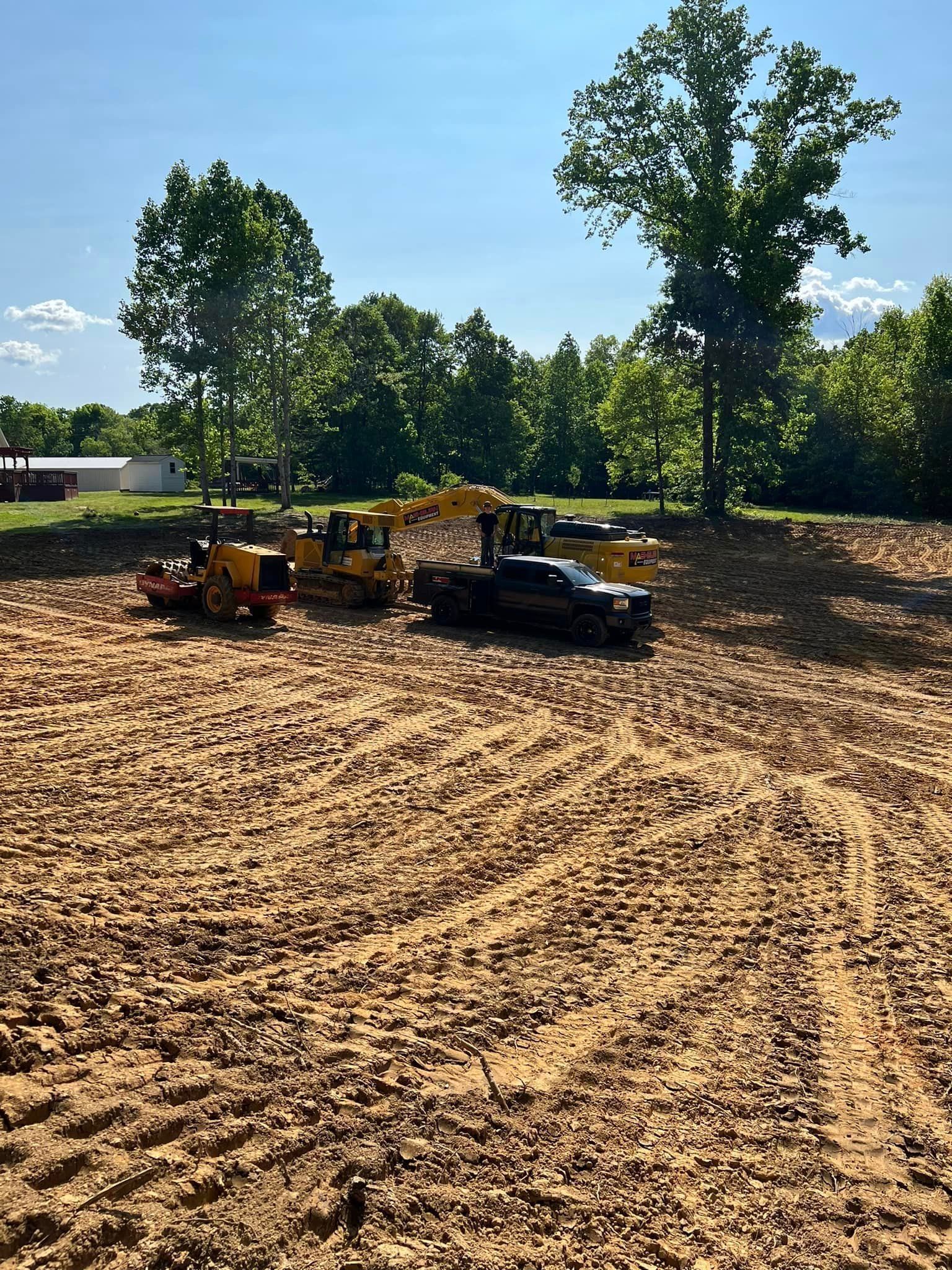Dirt lot with construction equipment and a pickup truck under a blue sky, trees in the background.