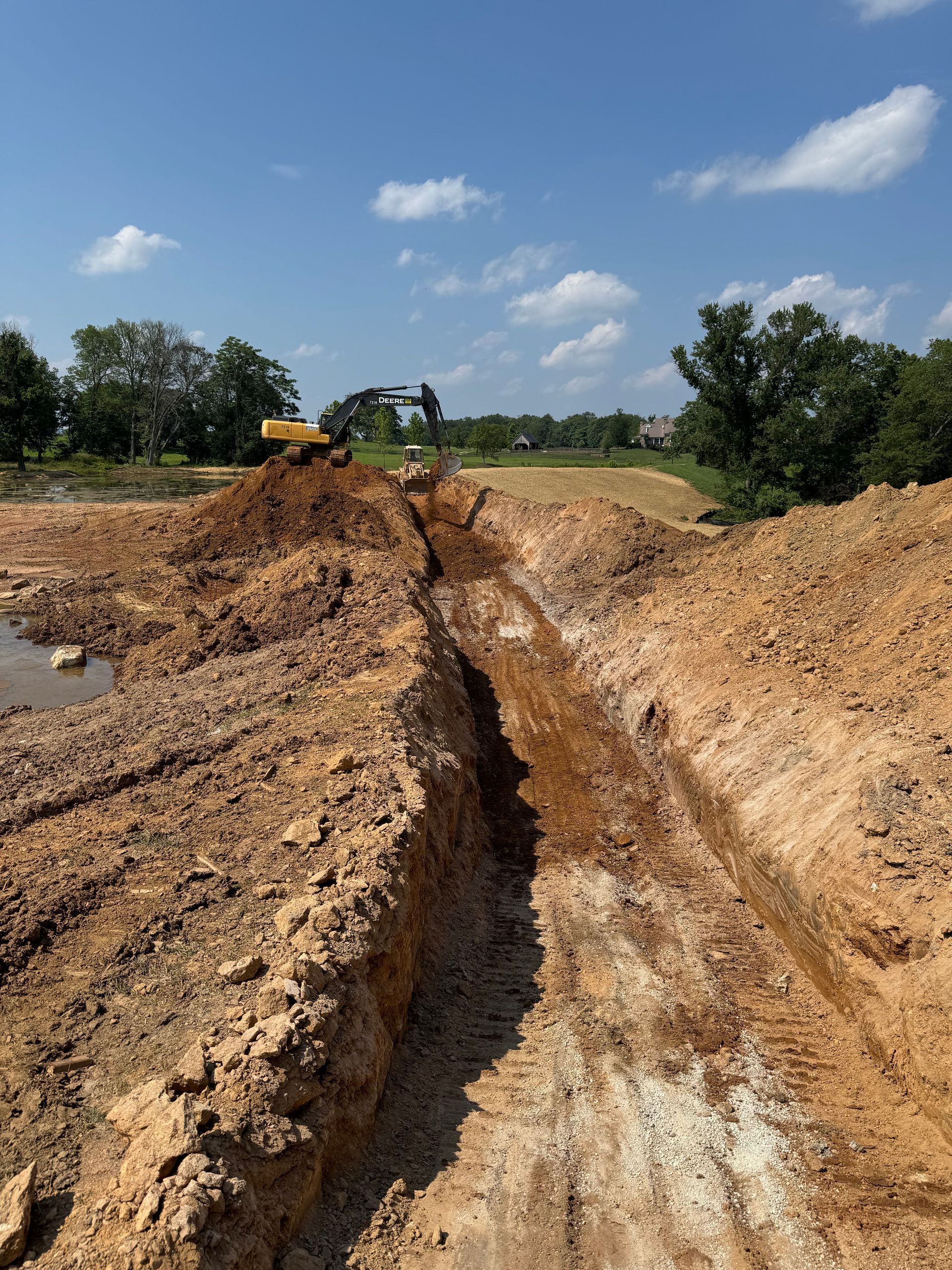 An excavator digs a trench in muddy, brown earth under a blue sky, near trees.