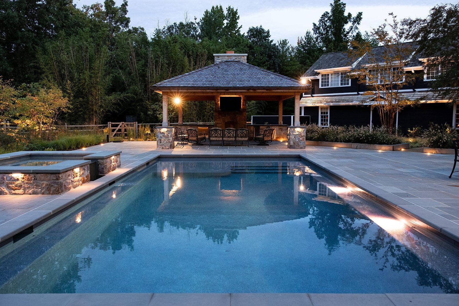 Outdoor pool at dusk beside a stone patio and lit pavilion, with trees in the background