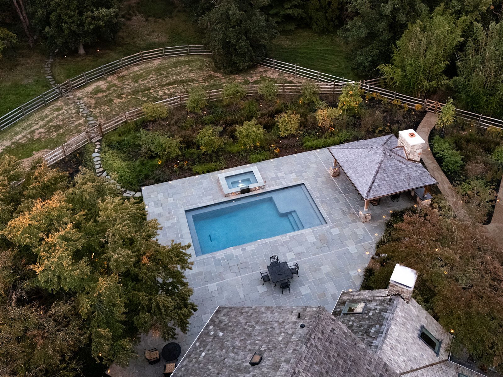 Aerial view of a backyard patio with a rectangular pool, hot tub, and shaded seating area surrounded by trees.