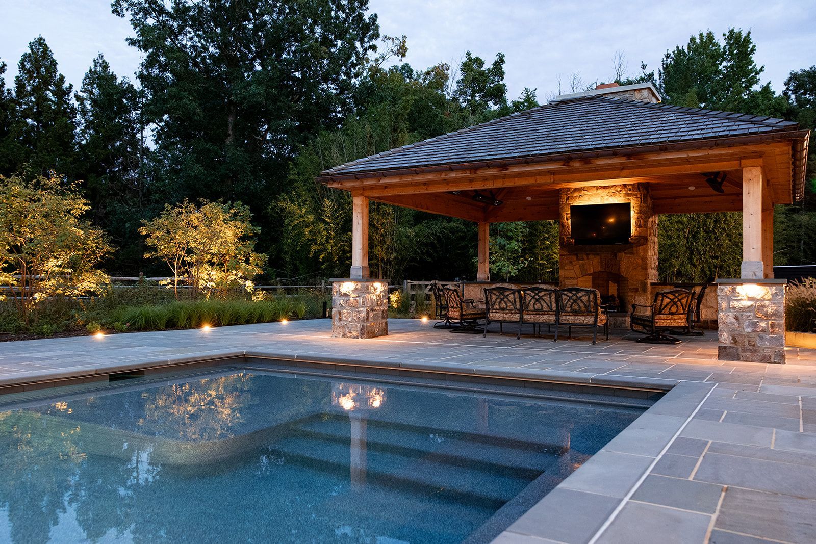 Stone patio pool beside a lit pavilion at dusk with trees in the background