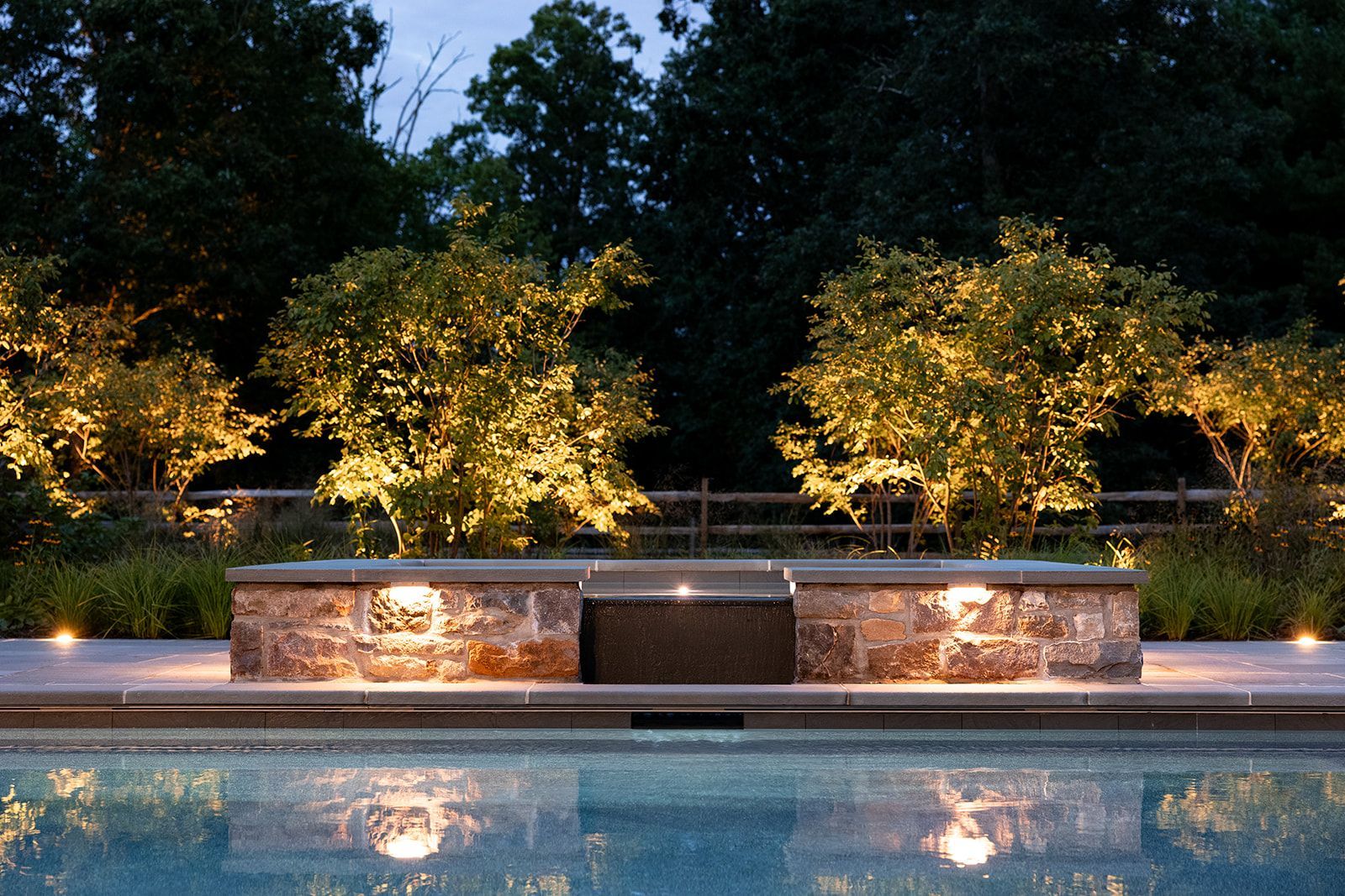 Illuminated backyard pool with stone water feature and lit trees at night