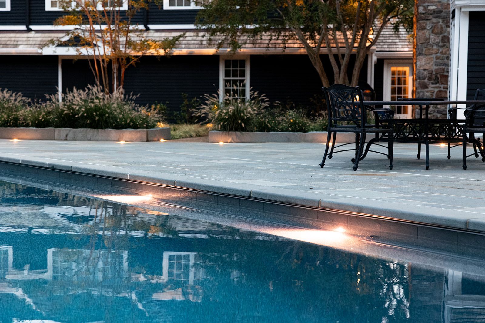 Poolside patio with blue water, black chairs, and warm lights beside a building at dusk