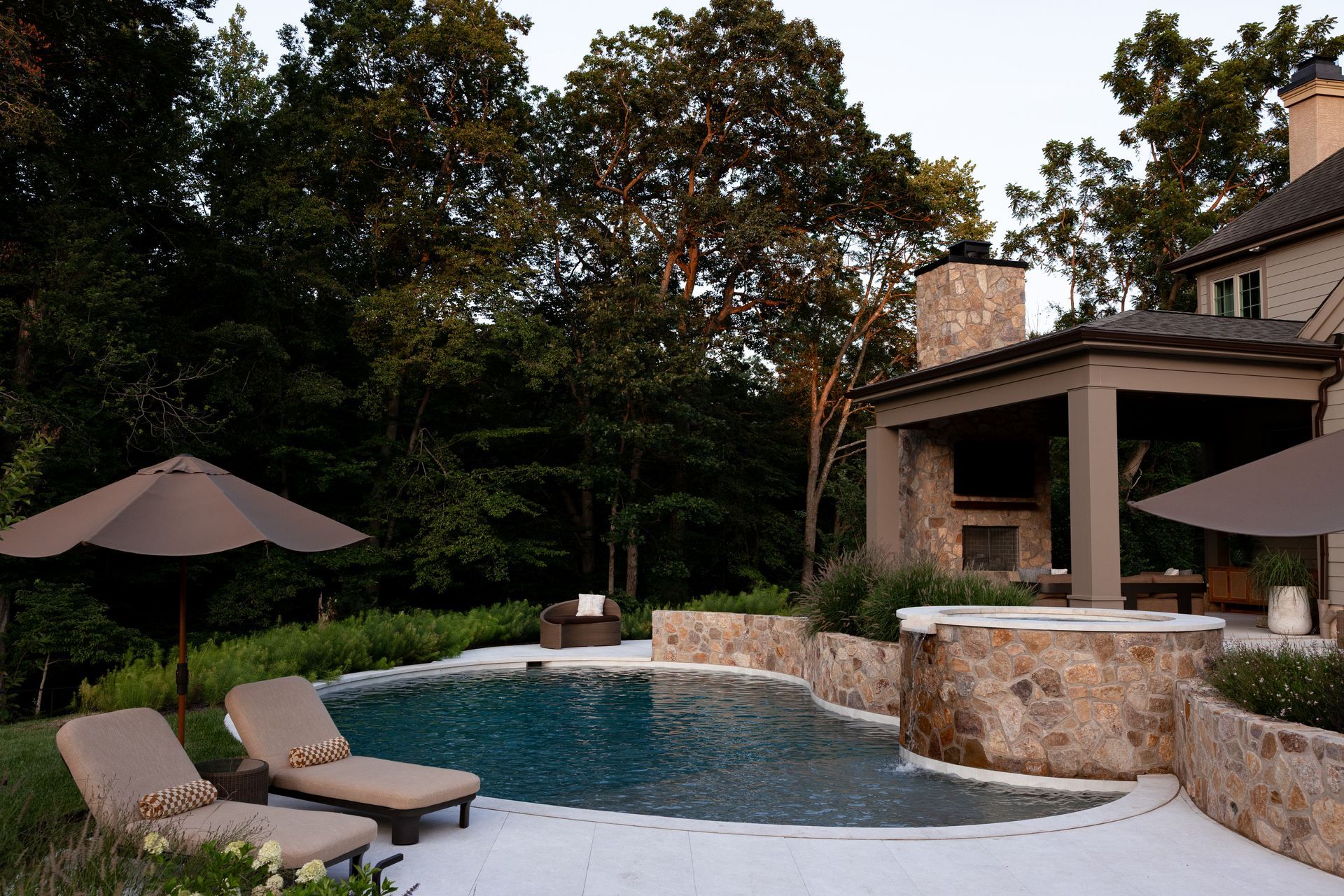 Poolside patio with pool, lounge chairs, umbrella, outdoor fireplace, and surrounding trees.
