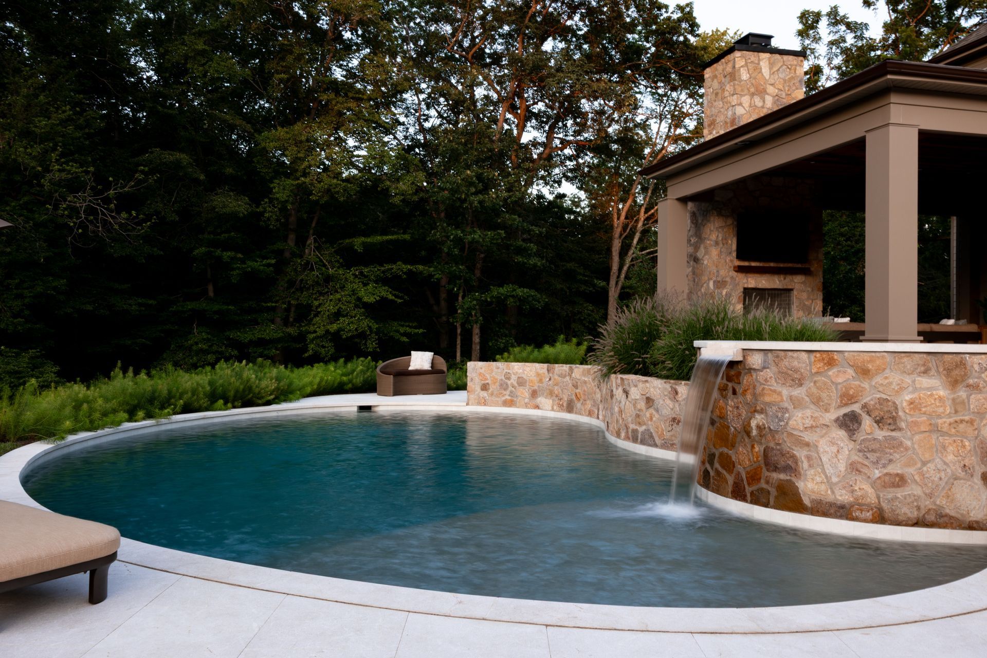 Pool with stone waterfall, surrounded by trees and a home's outdoor space.
