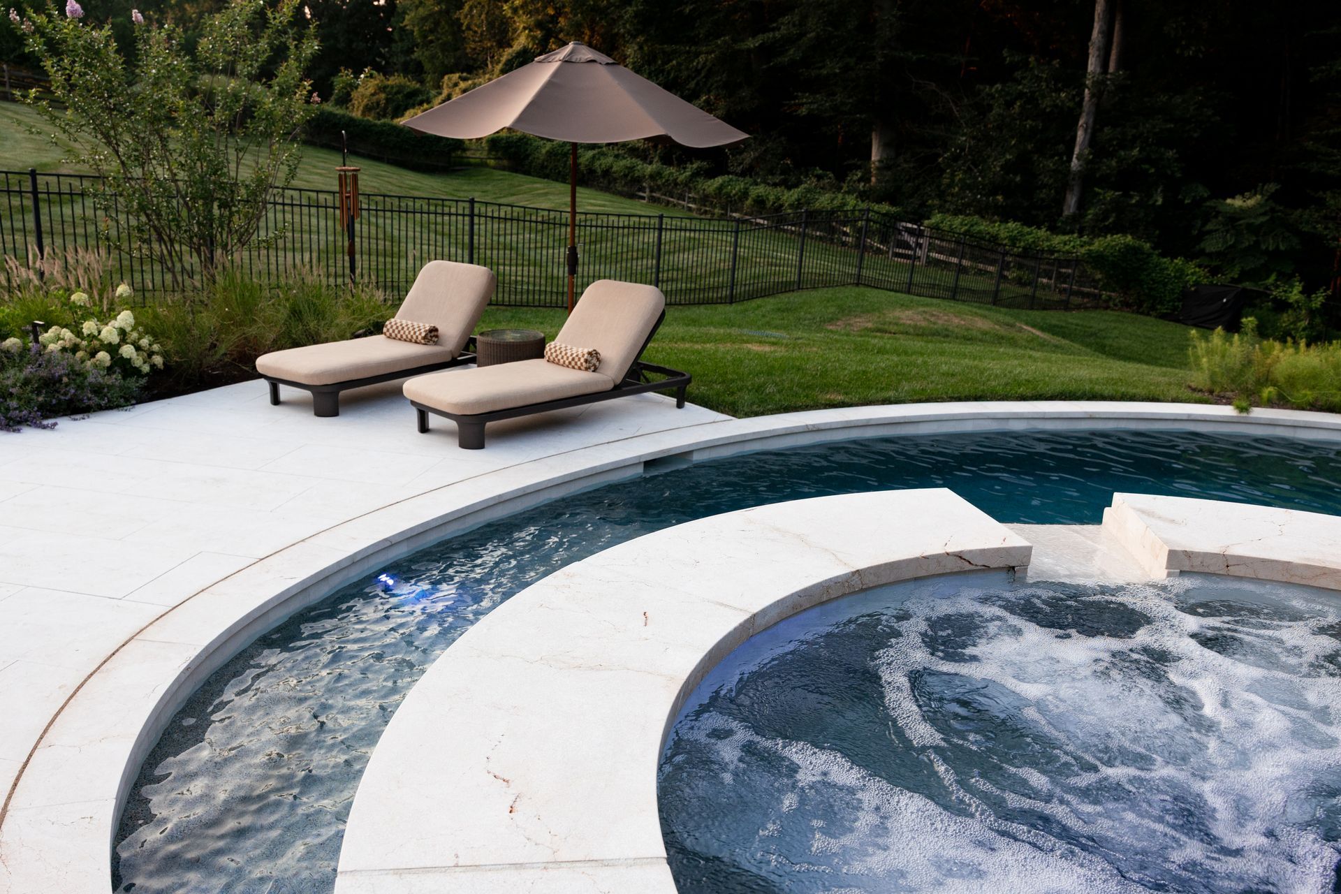 Pool with lounge chairs and umbrella on a patio, overlooking a lawn with a fence.